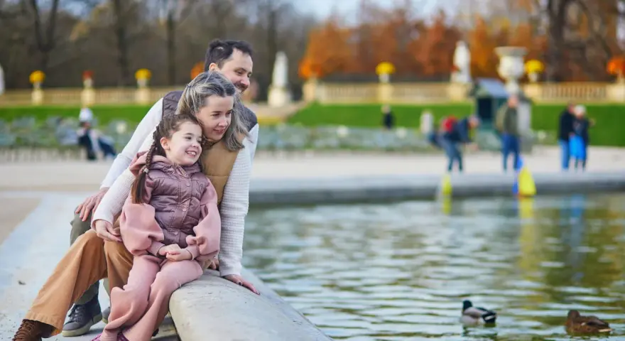Rodzina z dzieckiem siedzi nad brzegiem stawu z kaczkami w parku.