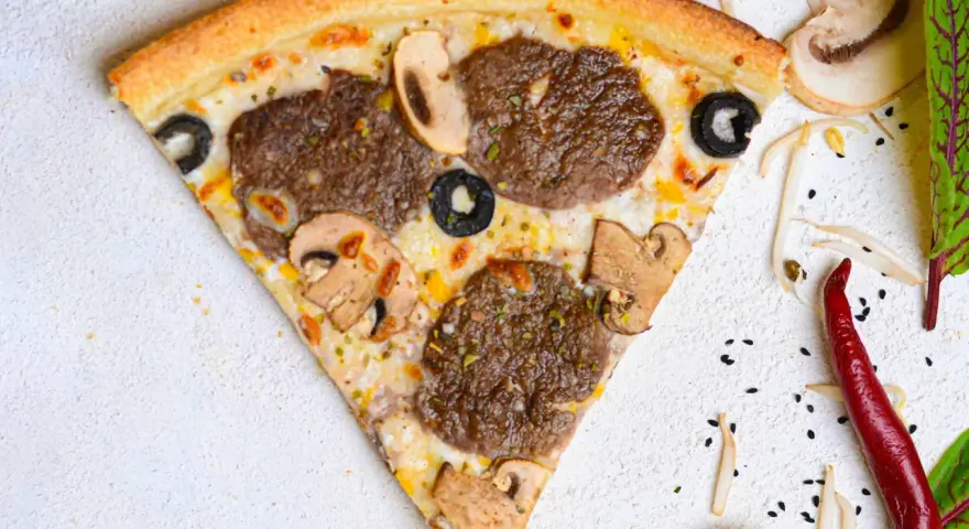 Kawałek pizzy z mięsem mielonym, pieczarkami i oliwkami na jasnym tle.