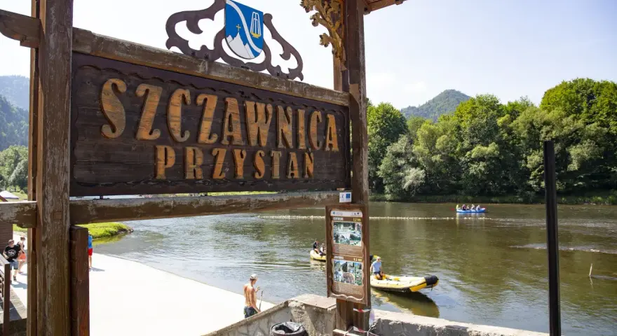 Drewniany szyld "Szczawnica Przystań" nad rzeką z tratwami i górami w tle.