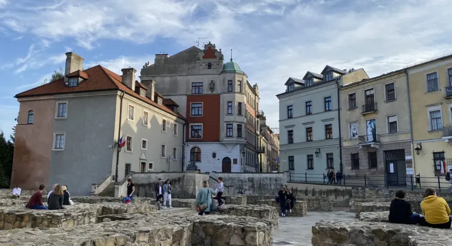 Ruiny zamku w Lublinie z ludźmi odpoczywającymi na kamiennych murach i zabytkowymi kamienicami w tle.