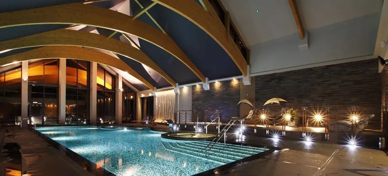 Nowoczesny hotel z basenem na Mazurach. Wnętrze z drewnianą konstrukcją dachu, kamienną ścianą i oświetleniem tworzącym relaksującą atmosferę.